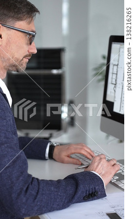 Architect designing building plans on computer, typing at desk with architectural model and blueprints. Business and architecture concept Architect designing building plans on computer, typing at desk with architectural model and blueprints. Business and architecture concept 138216265