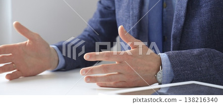 Professional businessman gesturing while discussing strategy, seated at white desk with tablet, eyeglasses, and wristwatch reflecting on sleek workspace surface. Business people concept Professional businessman gesturing while discussing strategy, seated at white desk with tablet, eyeglasses, and wristwatch reflecting on sleek workspace surface. Business people concept 138216340