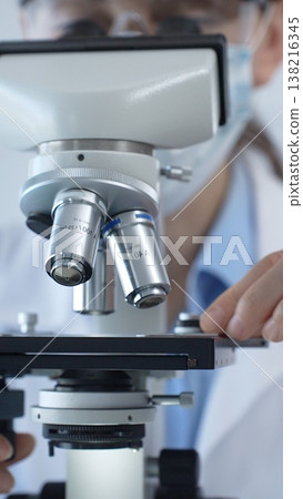 Female scientist wearing lab coat and protective glasses using a modern microscope in a laboratory setting, conducting research and experiments, close up. Health care, medicine and science concept Female scientist wearing lab coat and protective glasses using a modern microscope in a laboratory setting, conducting research and experiments, close up. Health care, medicine and science concept 138216345