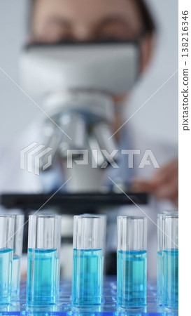 Close up of test tubes with blue liquid while scientist focusing on analyzing samples under a microscope in a laboratory setting. Medicine, healthcare and science concept 138216346