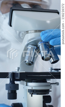 Close up of medical virologist in a hazmat suit, mask, and gloves, diligently working at a microscope in a laboratory researching viruses. Medicine and science concept Close up of medical virologist in a hazmat suit, mask, and gloves, diligently working at a microscope in a laboratory researching viruses. Medicine and science concept 138216372