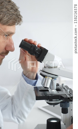 A man scientist carefully observing through a microscope in a brightly lit laboratory, conducting research for developing new medicines and treatments. Medicine and science concept 138216378
