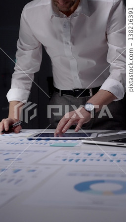 Professional man concentrating on important paperwork and statistics at his desk, working overtime in a dimly lit office environment during evening hours Professional man concentrating on important paperwork and statistics at his desk, working overtime in a dimly lit office environment during evening hours 138216391