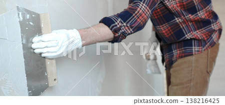 Unknown male construction worker wearing protective gloves smoothing plaster on interior wall during home renovation, applying material with professional trowel technique, close-up 138216425