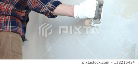 Unknown male construction worker wearing protective gloves smoothing plaster on interior wall during home renovation, applying material with professional trowel technique, close-up Unknown male construction worker wearing protective gloves smoothing plaster on interior wall during home renovation, applying material with professional trowel technique, close-up 138216454