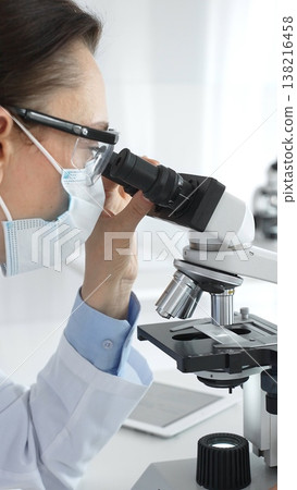 A female scientist in a protective mask and glasses carefully examines a sample under a microscope while conducting research in a modern laboratory. Concept of medicine and science 138216458