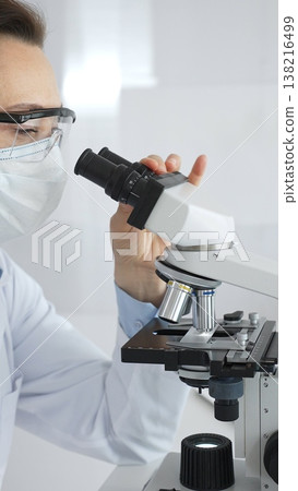 A female scientist in a protective mask and glasses carefully examines a sample under a microscope while conducting research in a modern laboratory. Concept of medicine and science 138216499