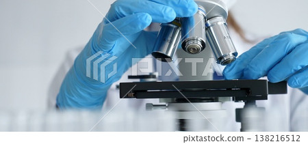 Close up of scientist's gloved hands adjusting microscope, conducting research in laboratory with test tubes containing blue liquid in background. Medicine, healthcare and science concept 138216512