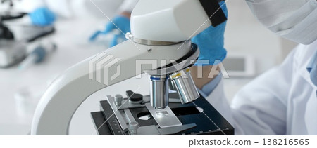 Scientific professional wearing blue protective gear examining microscope lens, performing detailed medical laboratory research. Medicine, healthcare and science concept 138216565