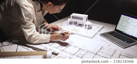 Architect carefully drafting blueprint plans on a desk with a building model and laptop, designing civil engineering projects and urban development Architect carefully drafting blueprint plans on a desk with a building model and laptop, designing civil engineering projects and urban development 138216566