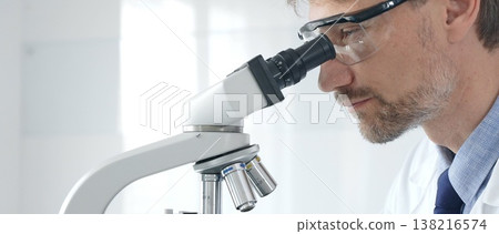 Adult male scientific researcher analyzing sample through microscope, wearing protective gear in high tech laboratory environment. Healthcare, medicine and science concept 138216574