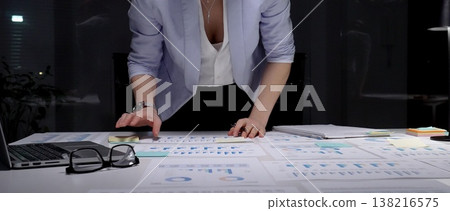 Businesswoman leaning over desk, working late in dark office, analyzing charts and graphs on paperwork, developing financial strategy and preparing for a deadline 138216575