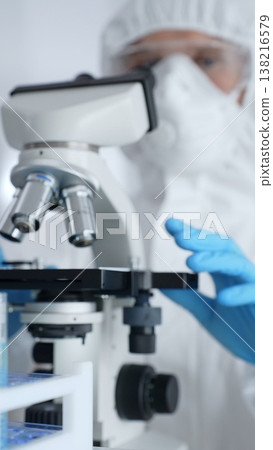Scientific researcher wearing protective gear adjusting microscope, analyzing lab samples with precision in sterile research environment. Medicine, healthcare and science concept 138216579