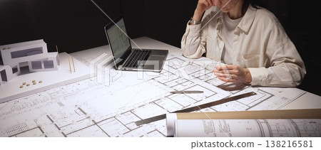 Female architect facing burnout after long late nights, rubbing tired eyes while holding glasses amid blueprints, laptop and a modern building model on her cluttered desk 138216581