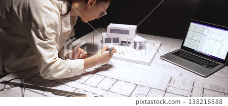 Architect carefully drafting blueprint plans on a desk with a building model and laptop, designing civil engineering projects and urban development 138216588