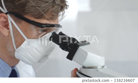 Male scientific researcher wearing protective gear examining sample through microscope, performing detailed laboratory investigation. Medicine and science concept 138216607