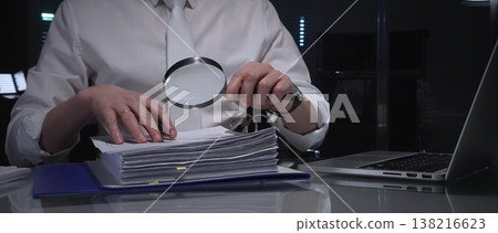 Businessman carefully examining financial documents with a magnifying glass, focusing on research and audit in a dark office. Audit and taxes in business 138216623
