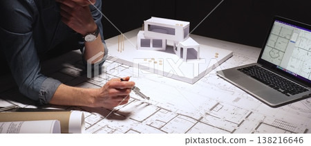 Architect drawing precise measurements and evaluating blueprints while also working on a physical architectural model and reviewing computer aided design plans on a laptop 138216646