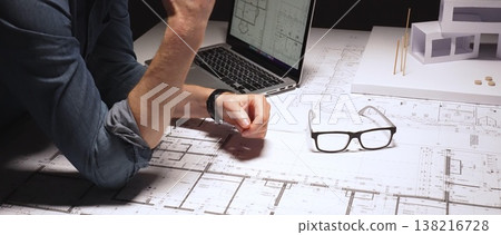 Architecture professional studying and working on construction blueprints and floor plans on a laptop computer, developing new projects with a physical model and design tools close by 138216728