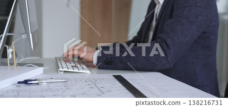 Professional architect or engineer typing on computer keyboard at office desk with building blueprints and design tools. Business and architecture concept Professional architect or engineer typing on computer keyboard at office desk with building blueprints and design tools. Business and architecture concept 138216731