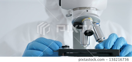 Close up of scientific researcher or virologist wearing protective gear is adjusting microscope, analyzing lab samples with precision in sterile research environment. Medicine and science concept 138216736