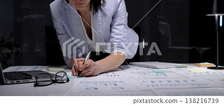 Businesswoman leaning over desk, working late in dark office, analyzing charts and graphs on paperwork, developing financial strategy and preparing for a deadline 138216739