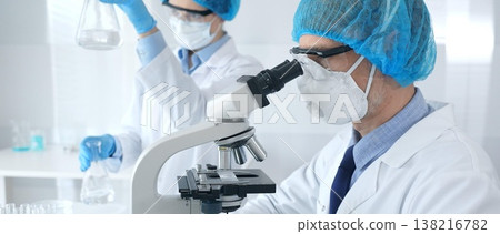 Masked male scientist in protective gear examining specimen under microscope while laboratory assistant handles glassware at the background. Medicine, science concept Masked male scientist in protective gear examining specimen under microscope while laboratory assistant handles glassware at the background. Medicine, science concept 138216782