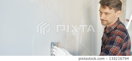 Construction worker man wearing gloves and holding a putty knife is applying plaster on a white wall, performing home renovation and improvement Construction worker man wearing gloves and holding a putty knife is applying plaster on a white wall, performing home renovation and improvement 138216794