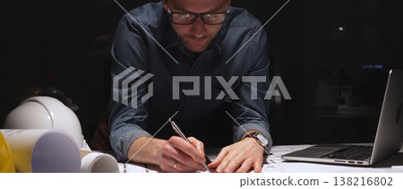 Architect or engineer working late at night in a dark office, concentrating on building blueprints, meticulously using a ruler for precise construction design, showing dedication and overtime work 138216802