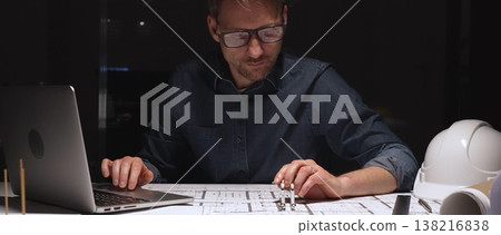 Architect or engineer working late, analyzing building blueprints and plans with a laptop, focusing on detailed architectural design during an intense night shift 138216838
