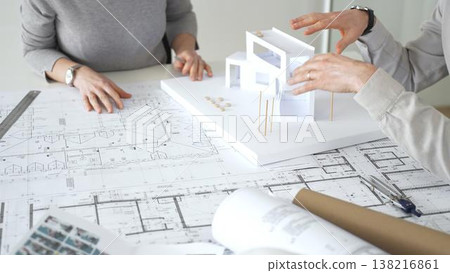 Architects collaborating, examining building blueprints with rulers and pens, developing innovative architectural designs and construction plans around a model house, close up Architects collaborating, examining building blueprints with rulers and pens, developing innovative architectural designs and construction plans around a model house, close up 138216861