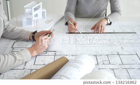 Architects collaborating, examining building blueprints with rulers and pens, developing innovative architectural designs and construction plans around a model house, close up 138216862