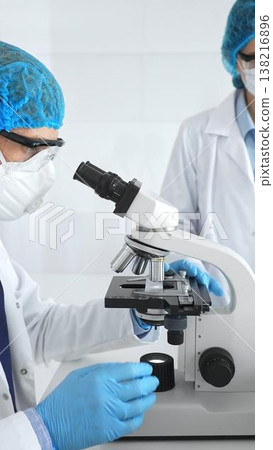 Two scientists, a man and a woman, in lab coats, masks and protective glasses conduct research with a microscope in a laboratory. Concept of medicine and science Two scientists, a man and a woman, in lab coats, masks and protective glasses conduct research with a microscope in a laboratory. Concept of medicine and science 138216896