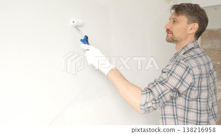 Man professional painter wearing checkered shirt, is rolling white paint on apartment wall with small blue roller, transforming interior space with steady strokes during comprehensive home renovation 138216958