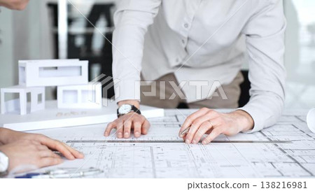 Architects collaborating over blueprints and a scaled model, discussing construction plans, urban development and design solutions in a modern office meeting setting, close up Architects collaborating over blueprints and a scaled model, discussing construction plans, urban development and design solutions in a modern office meeting setting, close up 138216981