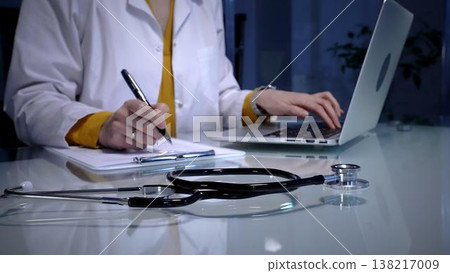 Stethoscope resting on a white reflective desk while medical professional working in clinic office. Wearing lab coat. Typing on laptop and writing notes. Stethoscope resting on a white reflective desk while medical professional working in clinic office. Wearing lab coat. Typing on laptop and writing notes. 138217009