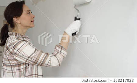 Middle aged woman wearing gloves is skillfully applying paint to a white wall with a paint roller in an apartment undergoing renovation, transforming the space with her creative touch Middle aged woman wearing gloves is skillfully applying paint to a white wall with a paint roller in an apartment undergoing renovation, transforming the space with her creative touch 138217028