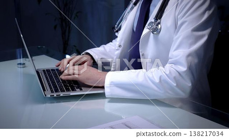 Medical doctor in lab coat with stethoscope typing on laptop in a dim office, managing electronic health records and telemedicine data for modern digital healthcare workflows 138217034