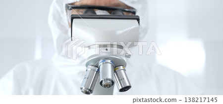 Medical virologist in a hazmat suit, mask, and blue protective gloves, carefully working at a microscope in a laboratory researching viruses. Healthcare and science concept Medical virologist in a hazmat suit, mask, and blue protective gloves, carefully working at a microscope in a laboratory researching viruses. Healthcare and science concept 138217145