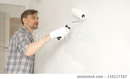 Man professional painter wearing checkered shirt, is rolling white paint on apartment wall with paint roller, transforming interior space with steady strokes during comprehensive home renovation 138217187