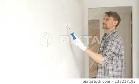 Man professional painter wearing checkered shirt, is rolling white paint on apartment wall, transforming interior space with steady strokes during comprehensive home renovation 138217192