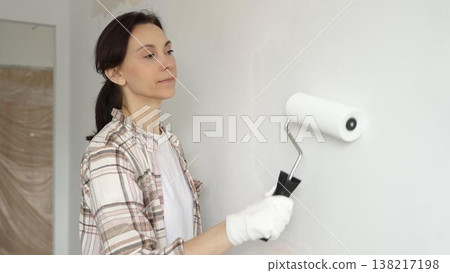 Middle aged woman wearing gloves is skillfully applying paint to a white wall with a paint roller in an apartment undergoing renovation, transforming the space with her creative touch Middle aged woman wearing gloves is skillfully applying paint to a white wall with a paint roller in an apartment undergoing renovation, transforming the space with her creative touch 138217198