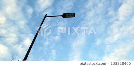 A modern LED street lamp stands tall against a bright, expansive blue sky filled with soft, white cirrocumulus clouds. The minimalist design of the black pole contrasts with the airy, natural backgrou 138218400