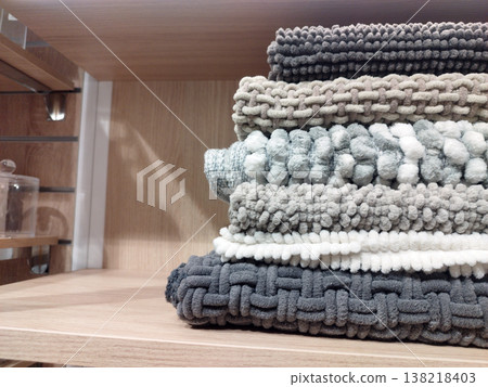 A stack of cozy, textured knit blankets in soothing shades of grey, beige, and white rests on a clean wooden shelf. The varied patterns, including chunky weaves and pom-poms, evoke a warm, homey feel 138218403