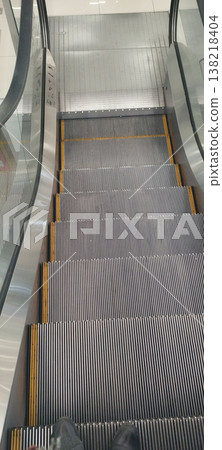 A high-angle, first-person perspective looking down a modern escalator in a bright, clean building. The metallic steps with yellow safety lines lead the eye toward the landing, reflecting a minimalist A high-angle, first-person perspective looking down a modern escalator in a bright, clean building. The metallic steps with yellow safety lines lead the eye toward the landing, reflecting a minimalist 138218404