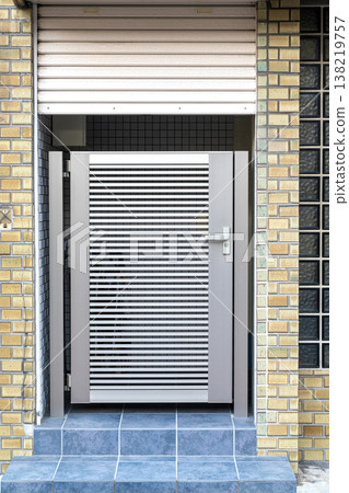 Modern silver metal gate with horizontal slats at a residential entrance. Tan brick walls, blue floor tiles, and a rolling shutter frame this clean contemporary urban architectural detail. Modern silver metal gate with horizontal slats at a residential entrance. Tan brick walls, blue floor tiles, and a rolling shutter frame this clean contemporary urban architectural detail. 138219757