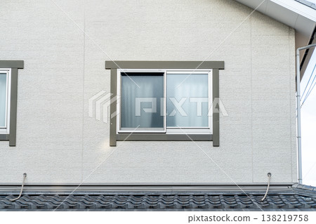 A close-up exterior view of a sliding frosted glass window on a modern residential house with beige textured walls and grey trim. Traditional tiled roof visible at the bottom. Japanese style house. A close-up exterior view of a sliding frosted glass window on a modern residential house with beige textured walls and grey trim. Traditional tiled roof visible at the bottom. Japanese style house. 138219758