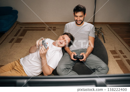 Two gay men relaxing at home, enjoying casual evening together, sharing comfortable moment while playing together video games with console controllers. Concept of modern online entertainment. Two gay men relaxing at home, enjoying casual evening together, sharing comfortable moment while playing together video games with console controllers. Concept of modern online entertainment. 138222400