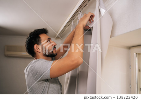 Low-angle view of young bearded man hanging curtains on rod during apartment cleaning, organizing home, performing household chores, doing routine house maintenance. 138222420