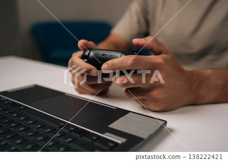Gamer hands holding black wireless gamepad, actively playing laptop video game sitting on white table at home, casual indoor gaming and leisure with modern portable technology. Gamer hands holding black wireless gamepad, actively playing laptop video game sitting on white table at home, casual indoor gaming and leisure with modern portable technology. 138222421
